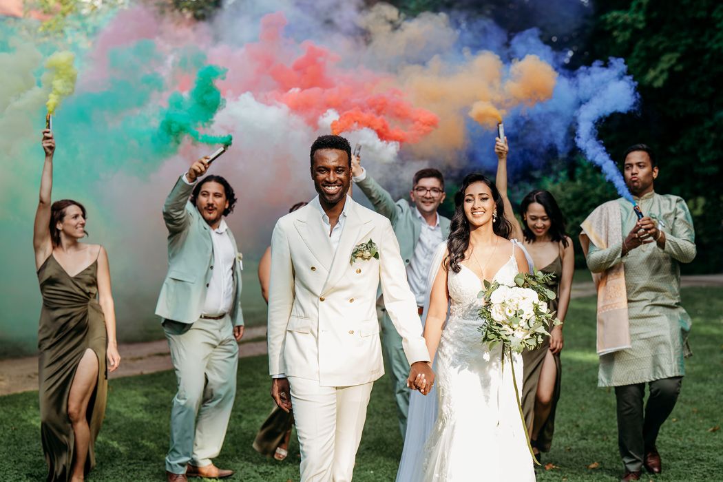 Brautpaar lächelt in die Kamera. Dahinter sind die Gäste mit bunten Rauchfackeln.