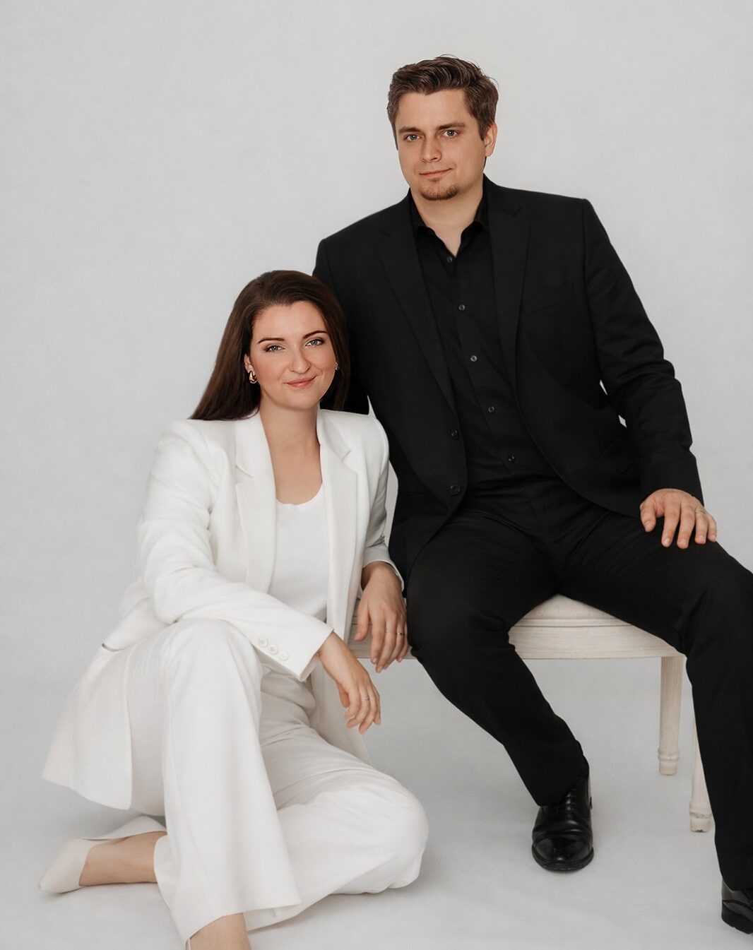 Patrick und Mariella Thanner Fotografenduo aus Baden. Sie sitzt lässig in weißen Hosenanzug am Boden. Er auf einem Hocker im dunklen Anzug.