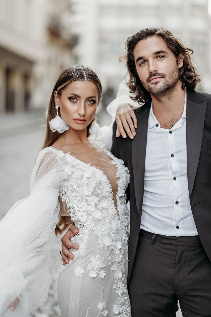 Ein Brautpaar posiert elegant in einer Stadt, wobei sie ein modernes Spitzenkleid mit tiefem Ausschnitt und auffälligem Haarschmuck trägt und er einen dunklen Anzug mit offenem weißem Hemd kombiniert.